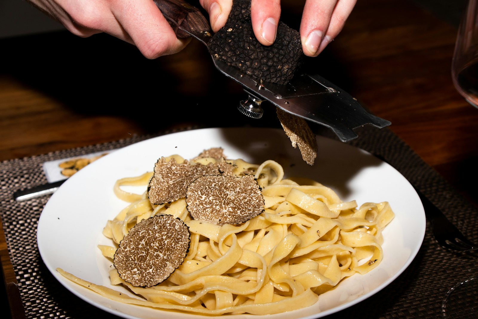 A close-up overhead view of fresh white truffle being shaved with a specialised slicer over a plate of fresh egg tagliatelle pasta on a wooden table, with the curls of pale truffle visible against the yellow pasta.